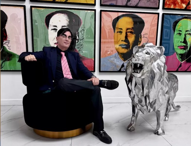 photo of a seated man with a conspicuous toupee beside a large lion figurine with ostensible Andy Warhol works in the background