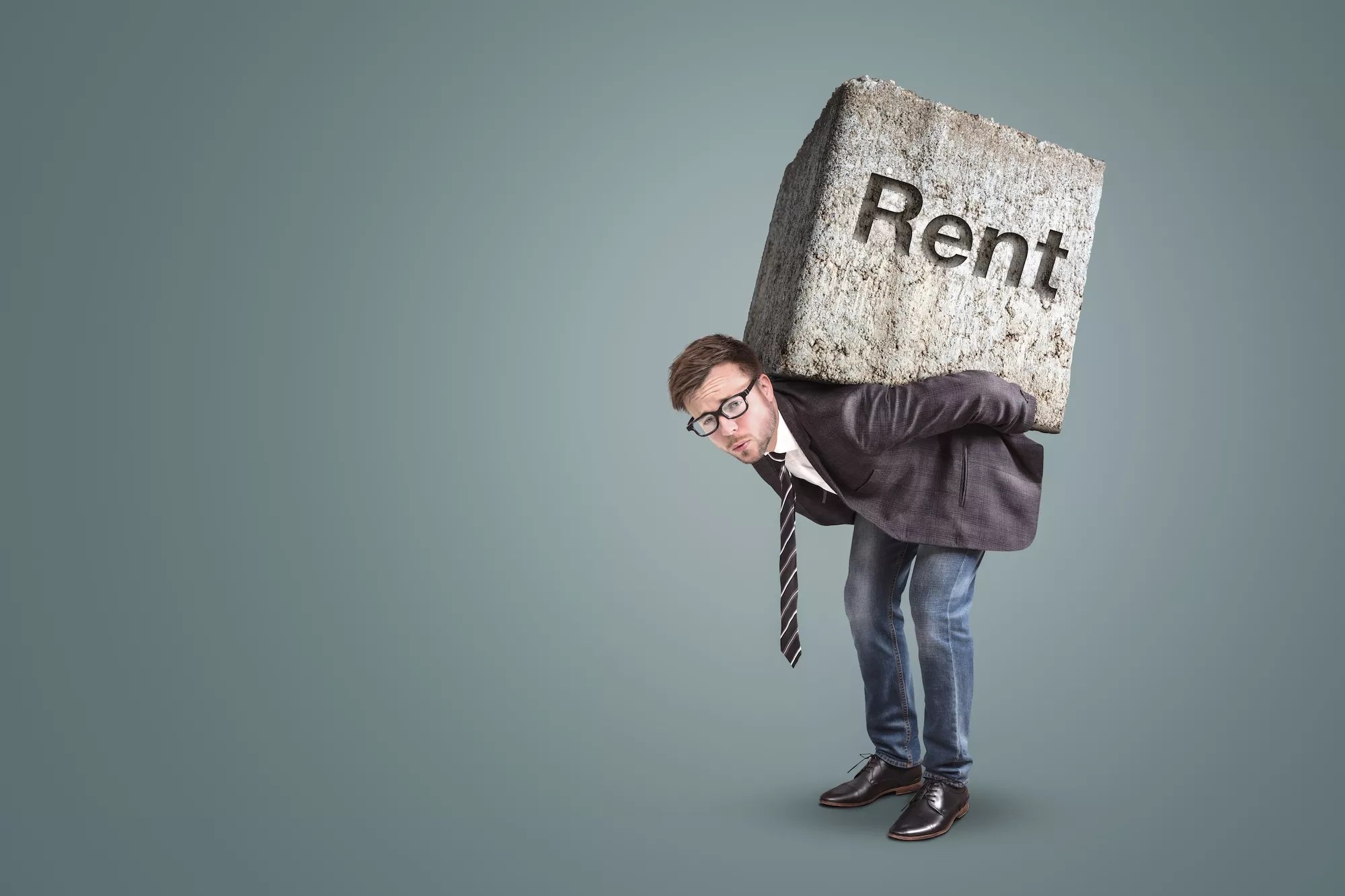 well-dressed hunched man with what looks to be a very heavy concrete block on his back with the word "Rent" etched into it  (Conceptual image of a man burdened by high rent)