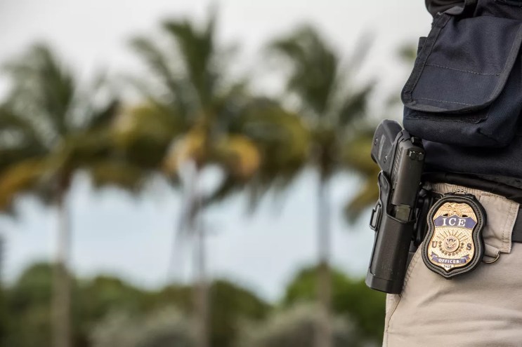 An immigration officer with an ICE badge next to palm trees.