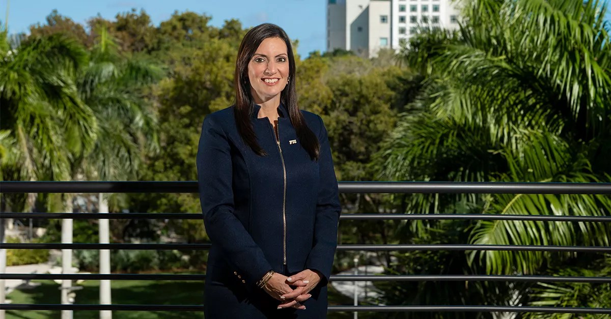 photo of a woman dressed in navy blue, standing with her hands clasped in front of her with tropical greenery and an FIU highrise in the background