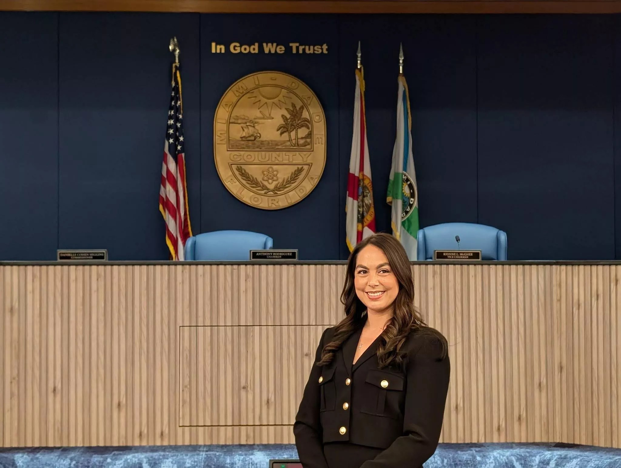 A brunette woman stands before a Miami-Dade County crest while wearing a black jacket