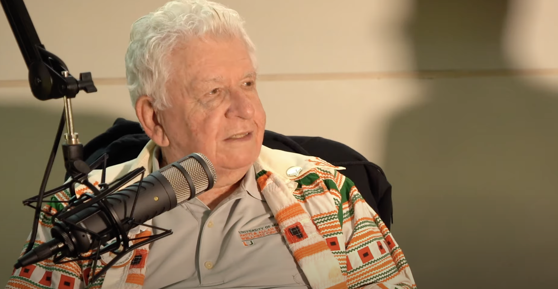 An elderly man with white hair looks to the side in front of a podcast microphone