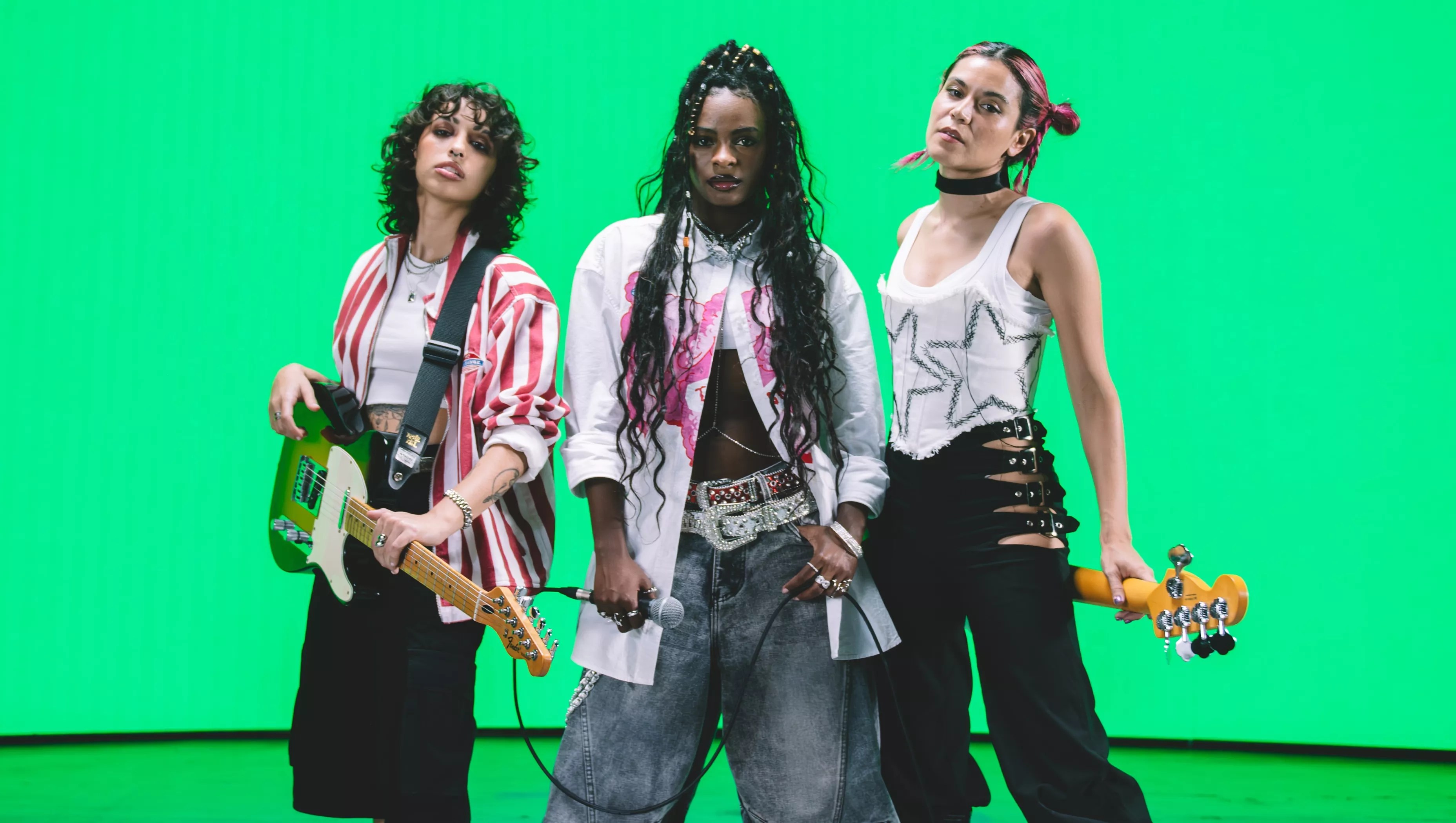 Three musicians pose in front of a bright green background. The woman on the left holds a guitar and the woman on the right holds a bass behind her back.