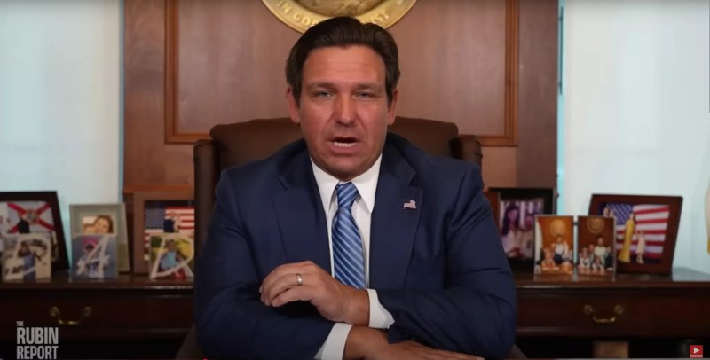 Ron DeSantis wearing a blue tie sitting at his desk