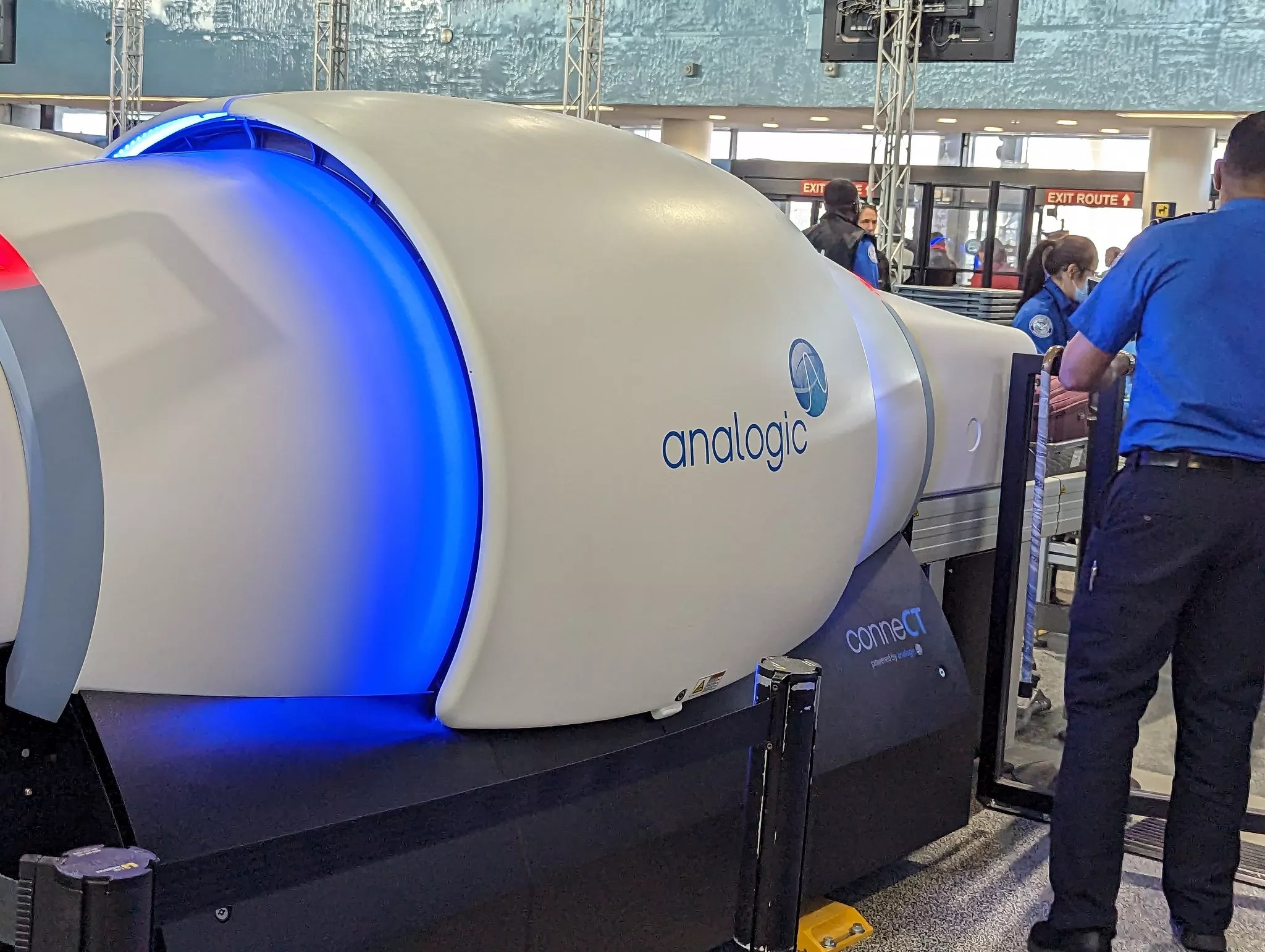 TSA agents surround a security scanning machine. A logo on the machine reads, "Analogic."