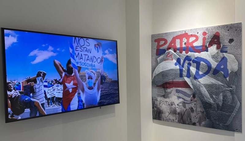 A TV in a museum displays images of Cuban protesters. One holds up a sign reading, "Nos Estan Matando (They're killing us) SOS Cuba." On the right of the TV, a painting displays the slogan, "Patria y Vida (Homeland and Life)"
