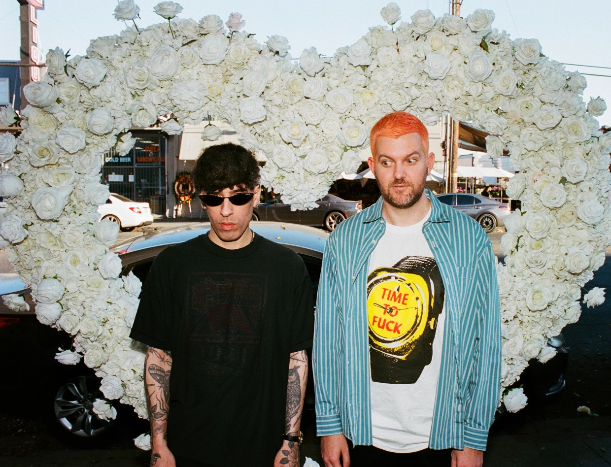 Two musicians are posing in front of a heart made out of flowers.