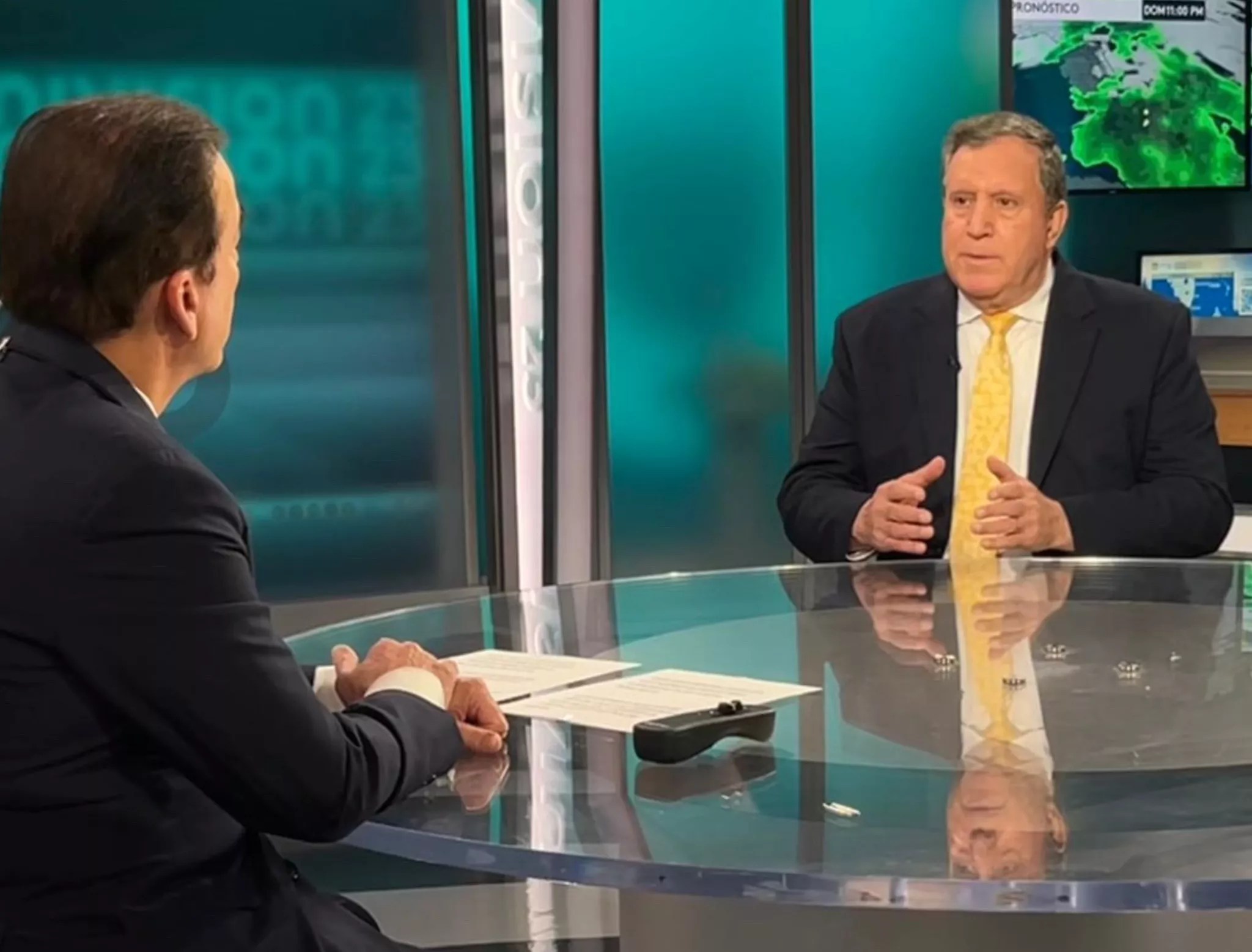 photo of a well-dressed man facing the camera, sitting at a glass table across from a television anchor who's partially visible in 3/4 profile