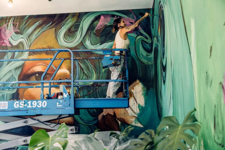 a woman stands on a platform to paint a mural
