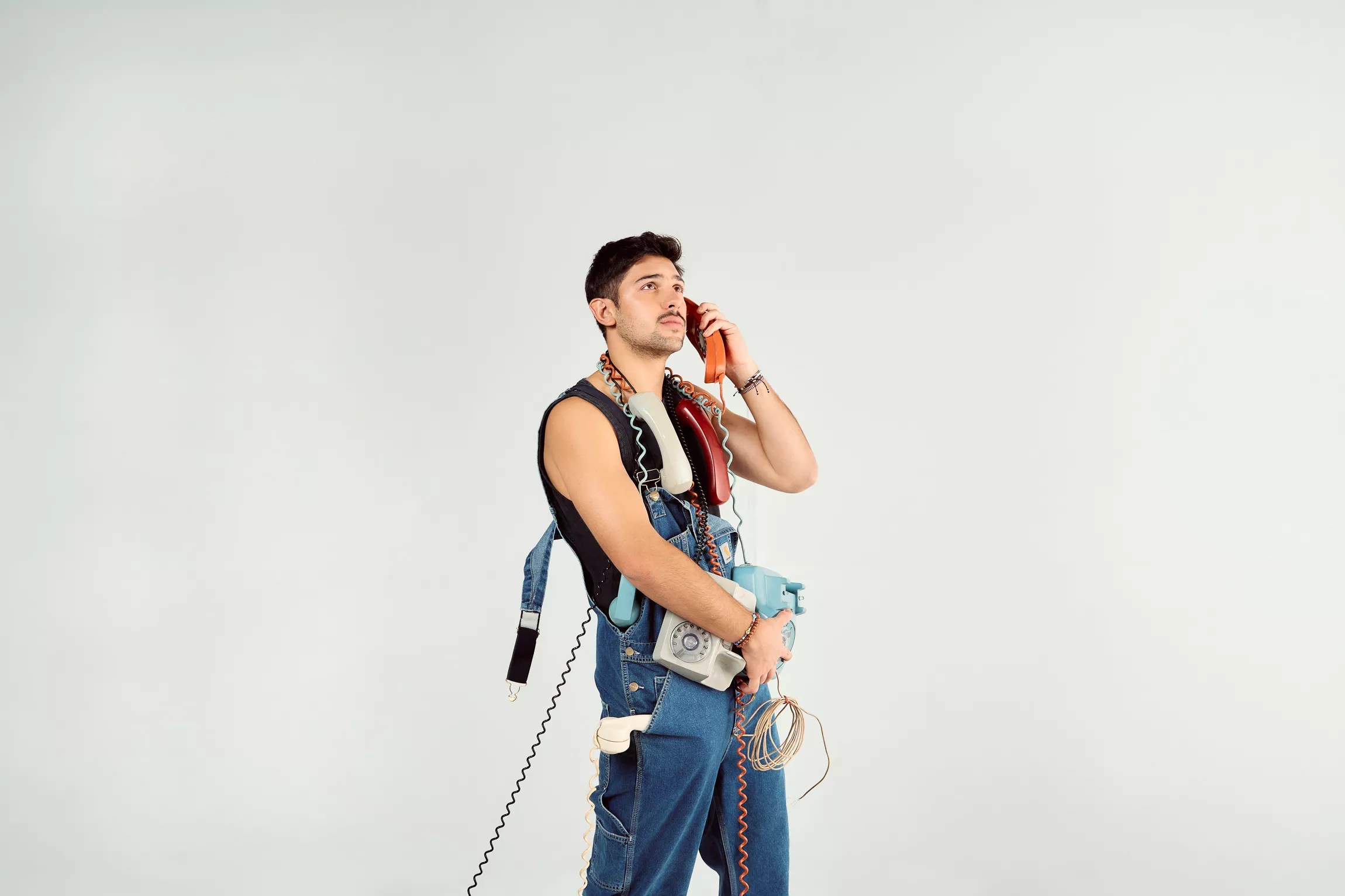 Portrait of a young man against a white backdrop holding many old telephones.