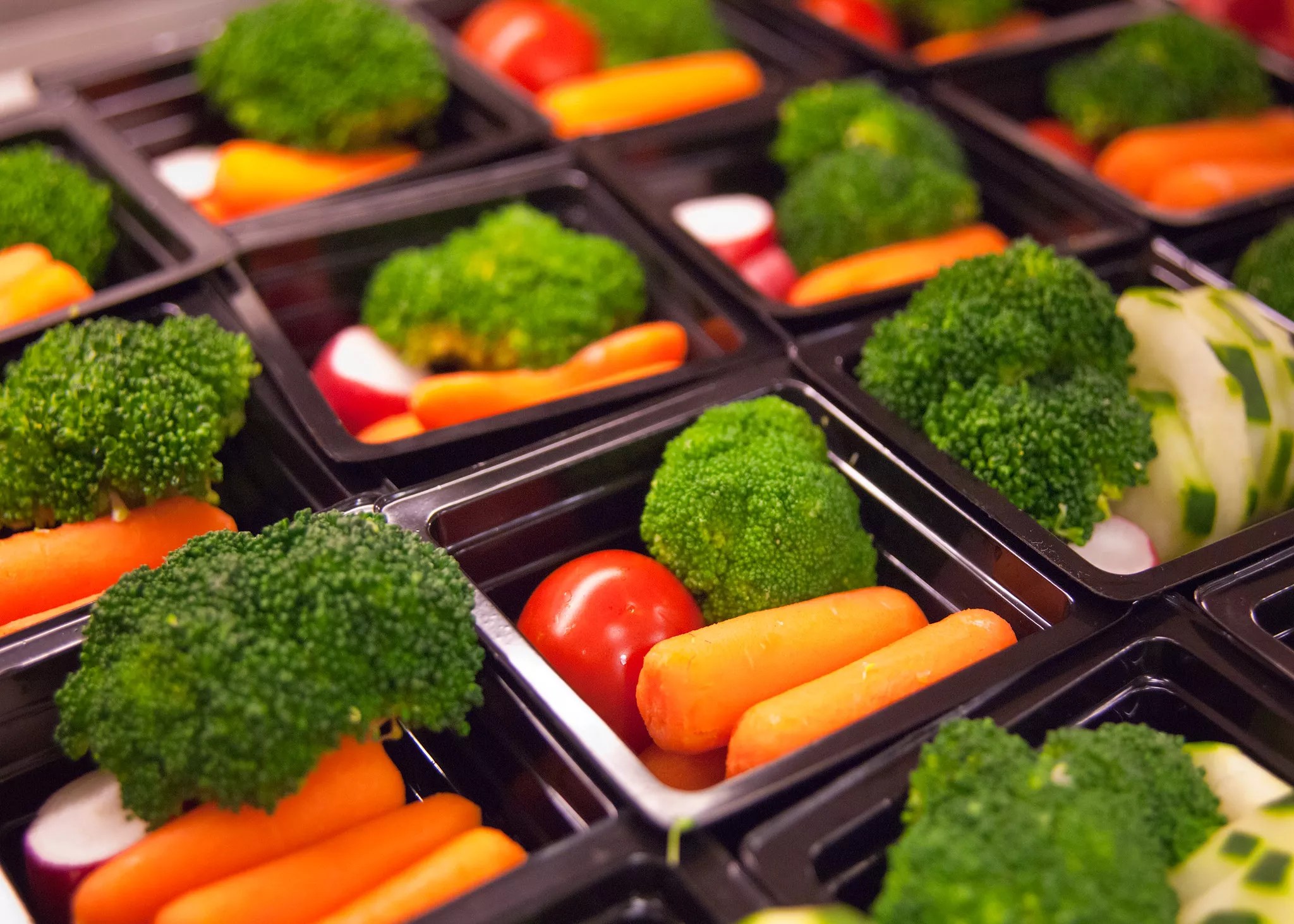Portions of vegetables like carrots, tomatoes, and broccoli in small black trays