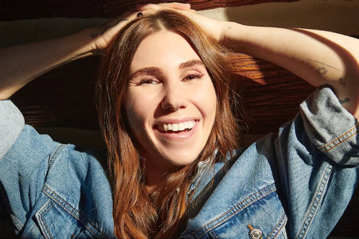 close-up portrait of actress Zosia Mamet wearing a denim jacket and resting her hands on her head