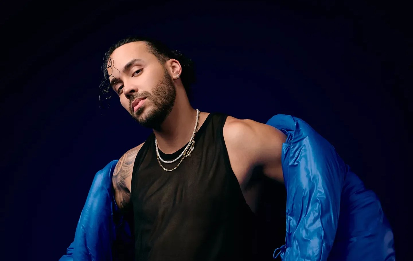 Portrait of a man posing for the camera wearing black pants, a black shirt, and a blue jacket.